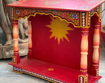 Puede incluir: Un pequeño estante de madera pintado a mano con una base roja y diseños florales amarillos, naranjas y verdes. El estante tiene un arco decorativo y un diseño de estrella en la parte posterior.