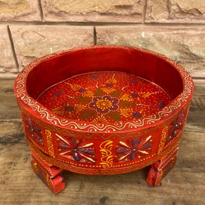 May include: A round, red, wooden decorative tray with a floral design on the inside. The tray has a raised edge and is supported by four short legs. The tray is painted with intricate patterns in yellow, blue, and white.