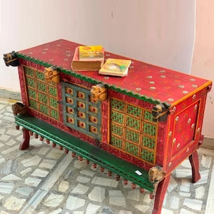 May include: A hand-painted wooden chest with intricate carvings and a green base. The chest has a red and green geometric pattern and a small door with a blue and green design. Two books are on top of the chest.