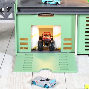 Op de afbeelding: Een groene speelgoedgarage met een rode monstertruck die door een autowasstraat rijdt. De garage heeft een helling en een grijs dak. Er staan ​​verschillende andere speelgoedauto's op de vloer.