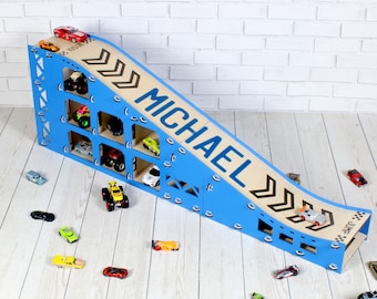Puede incluir: Rampa de coches de juguete de madera azul y marrón con un nombre personalizado "MICHAEL" en la rampa. La rampa tiene un área de almacenamiento para coches de juguete con varios estantes. La rampa está en un suelo de madera clara con varios coches de juguete esparcidos alrededor.