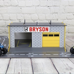May include: A wooden toy garage with a yellow door and a sign that says "BRYSON". The garage has a grey brick facade and a black roof. There is a yellow "PARKING AREA" sign inside the garage.