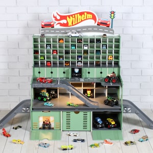Könnte beinhalten: Ein grünes Spielzeug-Garagenhaus aus Holz mit einer Rampe und mehreren Ebenen zum Verstauen von Spielzeugautos. Die Garage hat ein Schild mit der Aufschrift "Wilhelm" und ein rot-weiß-blaues Farbschema. Es befinden sich viele Spielzeugautos innerhalb und außerhalb der Garage.