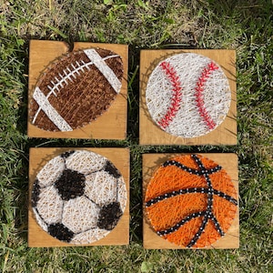 May include: Four wooden plaques with string art designs of different sports balls. The plaques are arranged in a row, with a football, baseball, soccer ball, and basketball. Each ball is made of white string with colored accents.