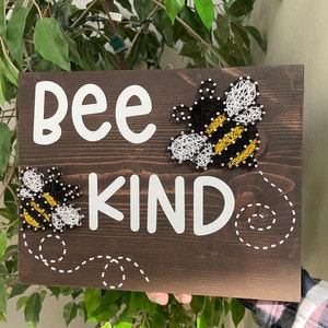 May include: A wooden sign with the words "Bee Kind" in white lettering. The sign features two string art bees, one on each side of the words. The bees are black, yellow, and white.