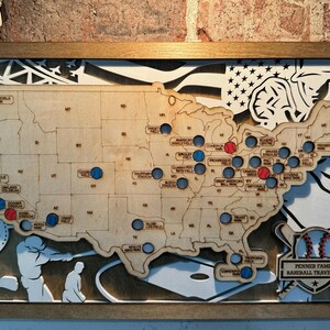 May include: A wooden map of the United States with blue and red dots marking locations. The map is framed in a brown wood frame and includes a baseball-themed design with the text "Penner Family Baseball Travel Map".