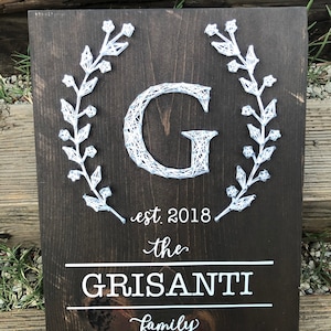 May include: A dark brown wooden sign with a white string art design of a letter G surrounded by a wreath. The sign also includes the text "est. 2018 the GRISANTI family".