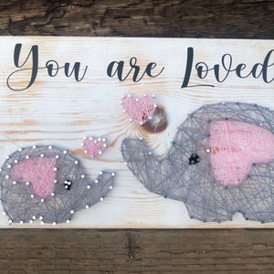 May include: A wooden sign with the text "You are Loved" in black cursive lettering. Two gray string art elephants with pink hearts are on the sign. The smaller elephant is on the left and the larger elephant is on the right.