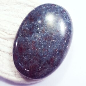 Może przedstawiać: Wypolerowany, owalny kamień szlachetny w odcieniach głębokiego niebiesko-szarego z czerwonobrązowymi wtrąceniami. Kamień ma błyszczącą, odbijającą światło powierzchnię i jest umieszczony na białym tle.
