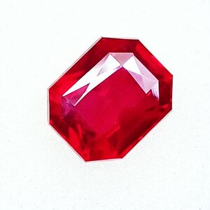 May include: A faceted, octagonal-shaped, ruby-red gemstone. The gem has a high-gloss finish and internal facets that reflect light. The gemstone is set against a white background, highlighting its vibrant color and cut.