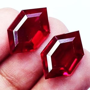 May include: Two loose, faceted gemstones with a deep red color and a distinct hexagonal shape. Each stone features sharp, geometric edges and a polished, reflective surface that catches the light, showcasing a vibrant, saturated crimson hue throughout.