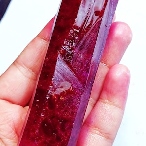 Peut inclure: Une pierre précieuse rubis rectangulaire et facettée, d'un rouge profond. La surface polie reflète la lumière, mettant en valeur la structure naturelle du cristal. La pierre est tenue dans une main, soulignant sa taille et sa forme.