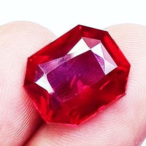 May include: A faceted, rectangular-cut ruby gemstone. The gem is a deep, saturated red with internal facets that reflect light. The gemstone is held in a person's hand.