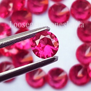 May include: A collection of round, faceted, vibrant pinkish-red gemstones. One gemstone is held in the centre by silver-toned metal tweezers, showcasing its circular cut and reflective surface. The text LoosegemstonesIndia is visible in white.
