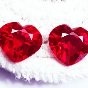 May include: Two vibrant red, heart-shaped faceted gemstones. Each stone features a classic heart cut with multiple triangular and kite-shaped facets that reflect light, creating a brilliant, sparkling appearance across the deep red surface.