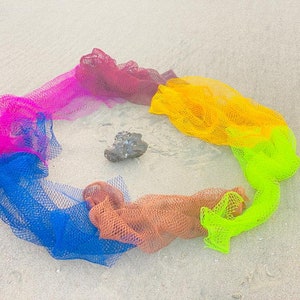 Pode incluir: Um círculo de rede colorida em uma praia de areia. A rede é em tons de rosa, vermelho, laranja, amarelo, verde e azul. Uma pequena pedra está no centro do círculo.