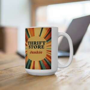 May include: White ceramic mug with a retro-style design featuring a sunburst pattern in shades of orange, yellow, green, and brown. The text "Thrift Store Junkie" is printed in black and red letters on the mug.