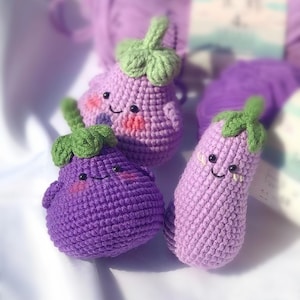 Puede incluir: Tres peluches de berenjena de crochet con caras sonrientes. Los peluches son morados y verdes y tienen una superficie texturizada.