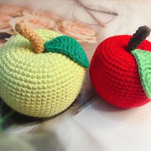 Può includere: Due giocattoli di frutta all'uncinetto, una mela verde e una mela rossa, con foglie verdi.