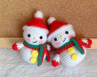 May include: Two crocheted snowmen wearing red Santa hats and green scarves. The snowmen are white with yellow buttons and black eyes.