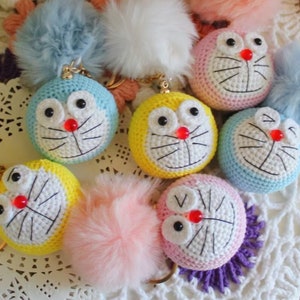 May include: A set of seven crocheted keychains featuring a cartoon cat character with different colored fur pom-poms. The keychains are made with white, blue, yellow, pink, and light blue yarn.