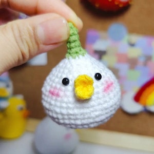 May include: A small crocheted chick with a white body, green stem, and yellow beak. The chick has black eyes and pink cheeks.