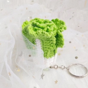 Puede incluir: Un llavero de ganchillo hecho a mano con forma de cogollo de lechuga verde y blanco. El llavero tiene un anillo y una cadena de metal plateado. La lechuga es de color verde vibrante, con blanco en la base.