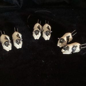 May include: A set of six ceramic corn holders, each shaped like a small cow with a white body and black spots. Each cow figure features two metal prongs extending from the back, designed for holding corn cobs securely during meals.