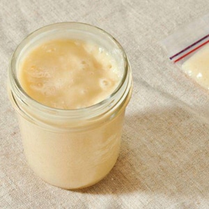 May include: A clear glass jar filled with a light beige, creamy substance. The jar is sitting on a beige fabric surface. A small plastic bag with a red and blue stripe is visible in the background, also containing a portion of the creamy substance.