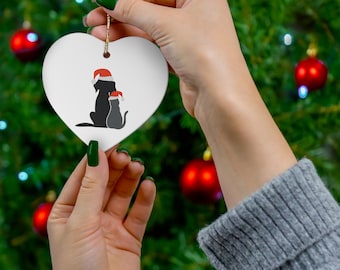 May include: A white heart-shaped ceramic ornament featuring a graphic illustration of a black dog and a gray cat sitting side by side, both wearing red and white Santa hats. The ornament is suspended by a thin gold string for hanging purposes.