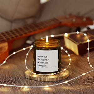Puede incluir: Una vela encendida en un tarro verde oscuro con una etiqueta blanca que dice "huele como lo contrario de las rodilleras de voleibol". La vela está sobre un posavasos de madera, rodeada de luces de cadena. Una guitarra y un libro están en el fondo.