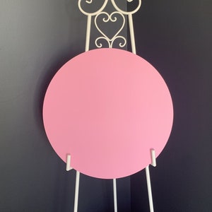 May include: A pink round sign on a white metal easel stand with decorative scrolls.