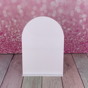 May include: A white, arched display stand with a flat base. The stand is made of a solid, opaque material and is set against a pink, glittery background. The stand is designed to hold or display items.