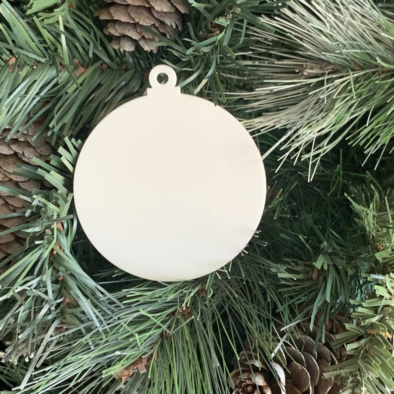 May include: A plain white wooden ornament in the shape of a circle with a small loop at the top. The ornament is hanging on a green Christmas tree.