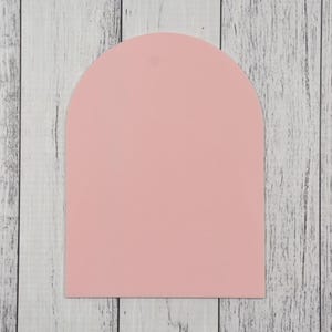 May include: A pale pink, arch-shaped sign with a small hole at the top. The sign is made of a smooth material and is set against a backdrop of white and grey wooden planks.