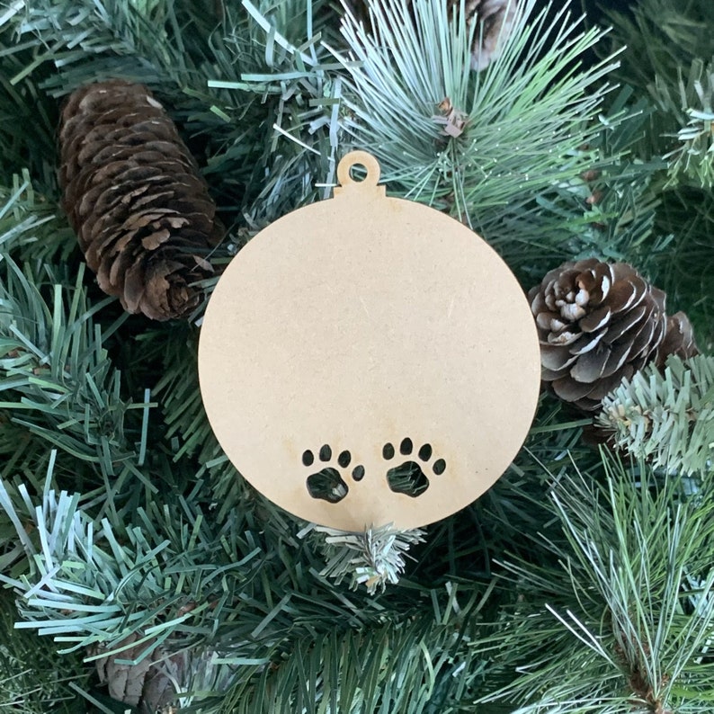 May include: A round, unfinished wooden ornament with two paw prints cut out. The ornament is hanging on a green Christmas tree.