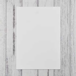 May include: A plain white rectangular sheet of paper, centrally positioned on a weathered, light grey wooden surface. The wood grain is visible, providing a textured backdrop to the smooth, unmarked paper. The composition is simple and clean.
