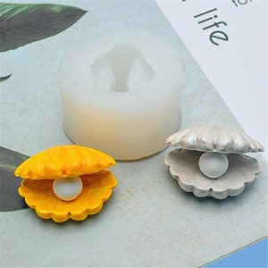 May include: Two silicone molds for making resin crafts. The molds are shaped like seashells with a pearl inside. One mold is yellow and the other is silver.