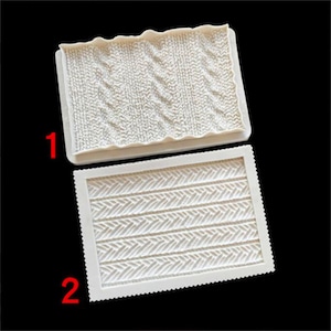 May include: Two white silicone moulds for crafting. The top mould features a cable knit pattern, while the bottom mould has a herringbone design. Both moulds are rectangular with decorative edges. Ideal for creating textured surfaces.