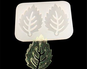 May include: A white silicone mold with two leaf shapes. The mold is used to create resin jewelry. A clear leaf with glitter is shown next to the mold.