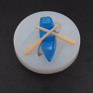 Op de afbeelding: Een witte siliconen mal met een blauw speelgoedbootje en twee houten peddels. De boot en peddels zijn over elkaar gekruist.