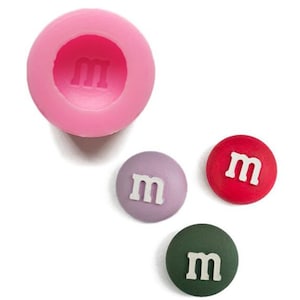 Könnte beinhalten: Rosa Silikonform mit einer einzigen Kavität in Form eines M&M-Bonbons. Drei M&M-Bonbons sind in verschiedenen Farben dargestellt: rot, lila und grün.