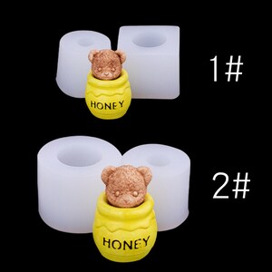 May include: Two white silicone molds for making resin crafts. Each mold features a bear figurine inside a honey pot with the word "Honey" printed on the pot. The molds are numbered 1 and 2.