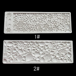 May include: Two white silicone moulds. One features a snowflake pattern and the other a seashell design. The moulds are rectangular and have the numbers "1#" and "2#" printed on them. These moulds are likely for crafting or baking.