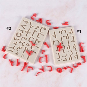 Peut inclure: Deux moules en silicone pour la cuisson, chacun avec un ensemble différent de lettres de l'alphabet arabe. Les moules sont blancs et les lettres sont en relief. Il y a aussi plusieurs lettres en plastique rouge éparpillées autour des moules.