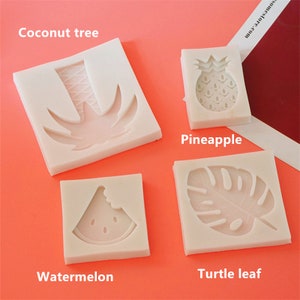 Peut inclure: Quatre moules en silicone blanc pour la cuisson ou l'artisanat. Les moules sont en forme de cocotier, d'ananas, de tranche de pastèque et de feuille de monstera. Les moules sont sur une surface rose.