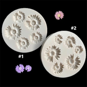 May include: Two white silicone molds for making flower-shaped decorations. The molds are numbered #1 and #2. Mold #1 has six flower shapes, while mold #2 has seven flower shapes.