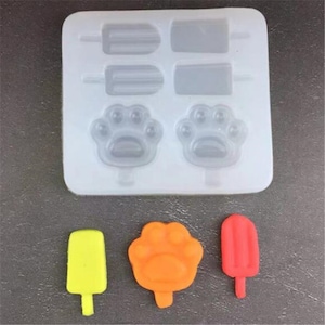 Może przedstawiać: Biała silikonowa forma z sześcioma wgłębieniami do robienia mrożonych przysmaków. Forma ma kształty lodów na patyku i śladów łap. Pokazano trzy gotowe przysmaki: żółty lód na patyku, pomarańczową łapę i czerwony lód na patyku.
