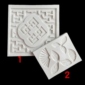 May include: Two white silicone moulds for baking. The first mould has a square shape with a geometric design in the centre. The second mould has a rectangular shape with three leaf-shaped designs.