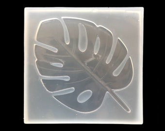 May include: A clear, square silicone mold featuring a detailed monstera leaf design. The leaf has several large, oval-shaped holes and prominent veins. The mold is designed for crafting and creating decorative elements.